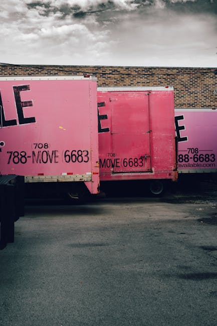 Several large pink moving containers with the logo and contact number of Man with Van Pratts Bottom are parked on a paved surface next to a brick wall under a cloudy sky. The containers are positioned side by side, with some doors closed and others slightly open, revealing interior space likely used for packing and transporting household items. The containers are part of a home relocation process, ready for furniture transport and loading onto a moving van. The scene suggests an exterior loading or unloading area as part of a professional removals service, with some visible equipment such as wheels and a trolley for handling large boxes and furniture. The overall setting emphasizes organized packing, careful handling, and logistics management involved in a structured moving operation, consistent with the services offered by Man with Van Pratts Bottom.