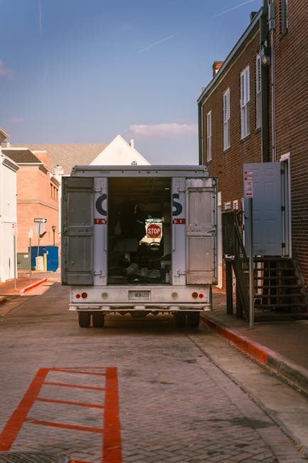 A large moving truck with open rear doors is parked on a narrow alleyway adjacent to a brick building, with moving boxes and wrapped furniture visible inside the vehicle. The truck is positioned near a set of external stairs leading to an upper floor of the building, and a stop sign is seen just behind the open doors. The area is illuminated by daylight, with clear blue sky overhead and nearby structures including other brick buildings and parked vehicles, indicating a home relocation or furniture transport process as part of a professional removals service provided by Man with Van Pratts Bottom.