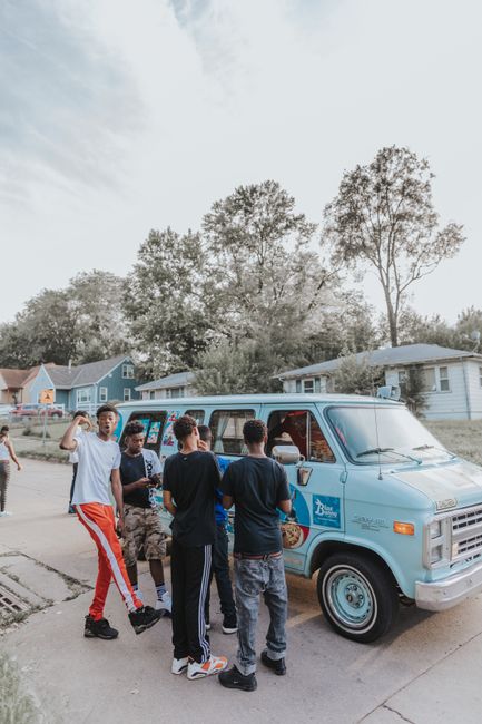 A group of five young men are gathered beside a light blue removal van, which is parked on a residential street with single-story houses and trees in the background. The men are engaged in conversation, with some facing the van and others turned towards each other. One individual is gesturing with his hand, while the others hold items such as small boxes or bags, suggesting they are in the process of packing or preparing for home relocation. The van has branding related to removals services, including a logo and text, and is positioned close to an open doorway of a house, indicating an ongoing loading process. The scene takes place during daytime with natural light illuminating the area, and the pavement is visible beneath the group's feet. This image depicts the initial stages of furniture transport and packing in a move managed by Man with Van Pratts Bottom, in line with home moving logistics and the preparation for furniture transport.