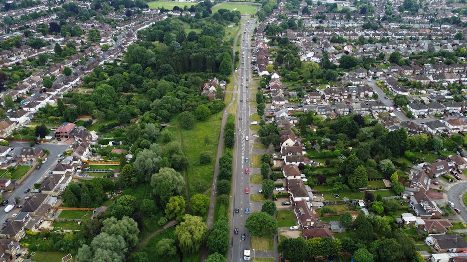An aerial view of a residential area in Pratts Bottom, Bromley, showing a straight street with parked cars along both sides, lined with detached and semi-detached houses featuring rooftops, gardens, and driveways. To the left of the street, there is a large green park area with dense trees, grass, and walking paths. On the right side, houses with various garden layouts are visible, some with fences, patios, and outdoor furniture. The scene is captured in daylight with clear skies, emphasizing the urban and suburban mix of properties. The image demonstrates typical elements involved in home relocation activities, such as packing boxes and furniture inside houses and preparing for furniture transport by [COMPANY_NAME], specialized in removals and moving services in the Bromley area. The road appears to be part of a logistical route used by the company's vans when carrying out packing and moving tasks in the neighbourhood.
