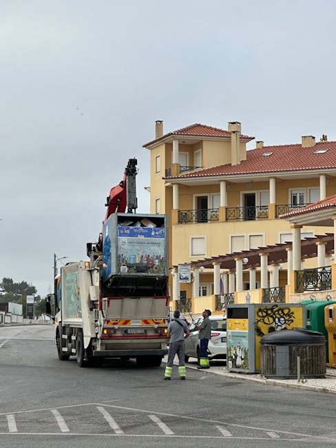 A large blue and white moving truck with a crane attached is parked on a street in front of a multi-story yellow residential building with balconies. The crane is extended upward and is lifting an object, possibly furniture or a large item, covered with plastic wrapping. Two workers dressed in high-visibility clothing stand nearby, observing the loading process. The truck is positioned near the pavement, and the building's entrance is visible in the background. Several objects, including a yellow recycling bin, a green waste bin, and a black trash bin, are situated on the sidewalk next to the truck. The scene takes place during daytime under overcast skies, with other vehicles and street furniture in the vicinity, illustrating a home relocation or furniture transport operation managed by Man with Van Pratts Bottom, experts in removals and moving services.
