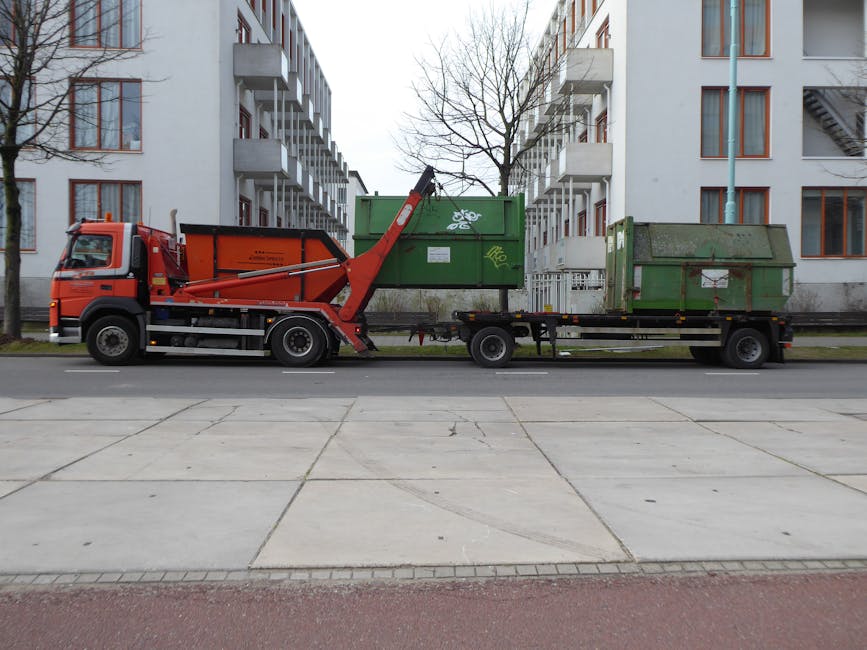 A red and black waste collection truck with a hydraulic lifting arm is parked on a residential street, adjacent to a sidewalk with large paving slabs. The truck is equipped for transporting bulky waste and is in the process of emptying a green recycling bin that has a white recycling symbol on its side, located near the cab. Behind the green bin, there is another green container with graffiti on it, positioned on an attached flatbed trailer. The scene is set against a background of mid-rise, white apartment buildings with multiple balconies and windows, and leafless trees indicating late autumn or winter. The truck is registered with Man with Van Pratts Bottom, a company specializing in removals, and the image reflects the logistics involved in home relocation, furniture transport, and packing and moving processes, illustrating professional waste disposal as part of property clearance or move preparation.