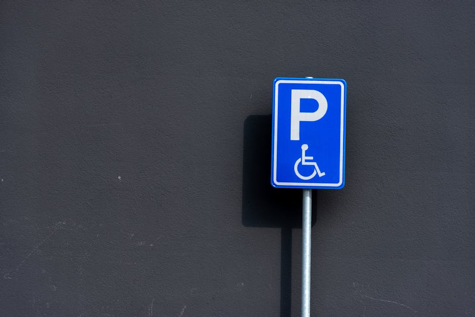 A blue and white parking permit sign indicating accessible parking for disabled individuals, mounted on a silver metal pole against a dark grey exterior wall. The sign features a large letter 'P' and a wheelchair symbol beneath it, with a subtle shadow cast on the wall. The setting appears to be outdoors, near a building entrance or parking area, which is relevant for property visits or home relocation services such as those provided by Man with Van Pratts Bottom. The sign's positioning suggests it is located on a pavement or driveway close to the property, supporting the logistics of loading or unloading furniture and moving boxes during a house removal or furniture transport operation. The clear visibility of the parking sign ensures proper parking arrangements during moving day, which is an important aspect for efficient planning in removals services.