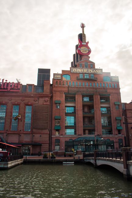A multi-storey brick building situated adjacent to a waterway, featuring several large blue-tinted windows and balconies. On the rooftop, there is a prominent neon sign shaped like a guitar with the words 'Music Hotel' and additional signage indicating 'Power Plant'. The building appears to be part of an entertainment or hospitality complex, with a cloudy sky overhead. The area in front of the building includes a paved walkway and a railing along the water's edge. The photograph captures the exterior during daylight hours, and the scene is relevant to house and furniture removals, with ample space for loading and unloading in preparation for home relocation or furniture transport services by companies like Man with Van Pratts Bottom.