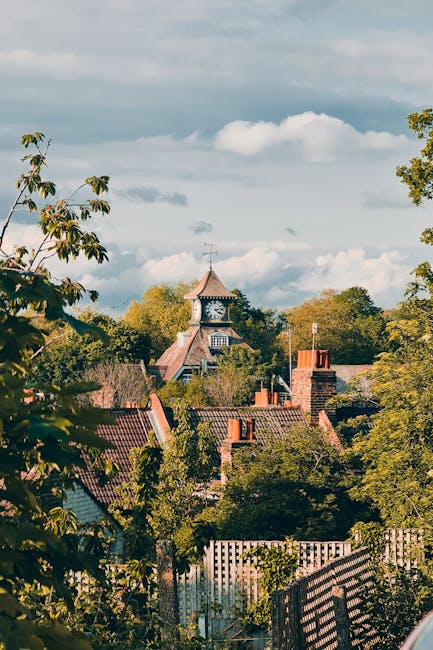 A view of a residential area during daytime with several rooftops and chimneys visible among lush green trees. In the background, a clock tower with a weather vane on top stands prominently above the surrounding houses. The sky is partly cloudy with patches of blue visible. This scene captures the tranquil atmosphere of a village setting, relevant to home relocation and house removals services offered by Man with Van Pratts Bottom, as seen on the Guide to Moving Near Pratts Bottom Village Green webpage.