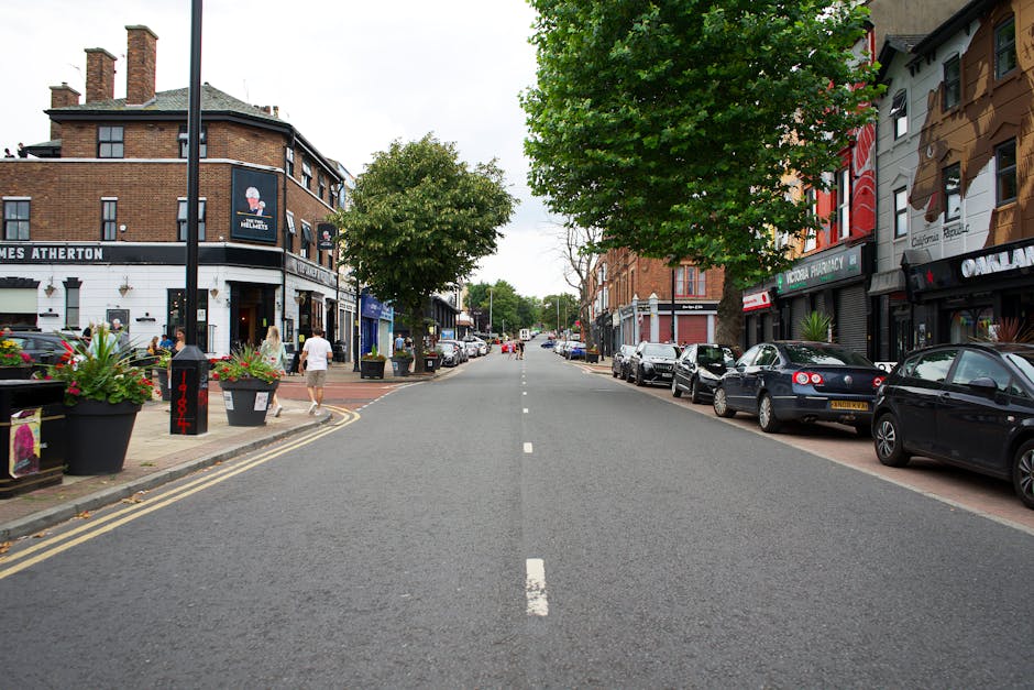 A quiet urban street scene near Pratts Bottom Village Green showing a row of parked cars along the right side of the road, including sedans and hatchbacks, with some visible ventilation grilles and vehicle registration plates. On the left pavement, there are large black flower pots filled with colorful flowers, and pedestrians are walking along the sidewalk, including a man in white shirt and beige trousers and a woman with long blonde hair. The street is lined with multi-story commercial buildings, including shops and cafes, with signage visible such as a pharmacy and other retail outlets. Large green trees with leafy canopies are positioned along the pavement, providing partial shade. Overhead, the sky is partly cloudy with soft lighting, and a van belonging to Man with Van Pratts Bottom is visible, suggesting ongoing home relocation or furniture transport activities nearby, framed by the street’s typical urban architecture and street furniture.