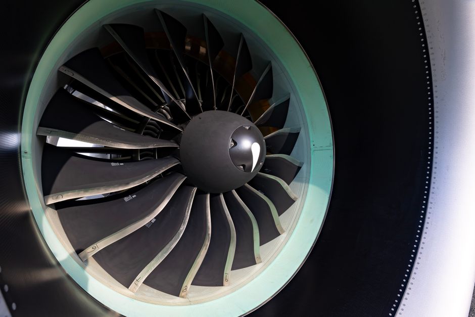 Close-up view of an aircraft jet engine's fan blades, showing multiple curved metal blades arranged in a circular pattern around the central hub. The blades are polished and reflective, with some ridg