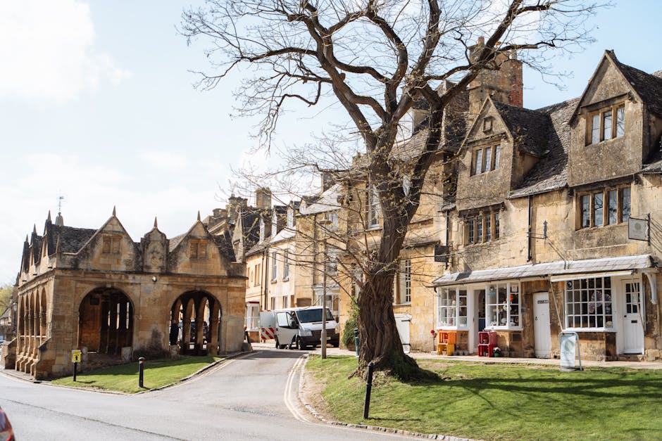 A street scene outside Pratts Bottom Village Hall during the home relocation process shows a stone-built structure with arched openings on the left side and a row of historic, beige-colored houses with pitched roofs and multiple windows on the right. In front of the houses, there is a leafless tree with sprawling branches and a well-maintained grassy area. Several vehicles, including a white van and a smaller delivery van, are parked close to the buildings, indicating the loading or unloading of furniture and packing materials. The scene is illuminated by natural daylight with a partly cloudy sky overhead, and some moving equipment such as dollies or straps may be seen in the vicinity, supporting the logistics of professional removals by Man with Van Pratts Bottom. This image captures the typical environment of a residential home relocation, where furniture transport and packing are coordinated within the local area, with the building exteriors and surrounding space clearly visible.