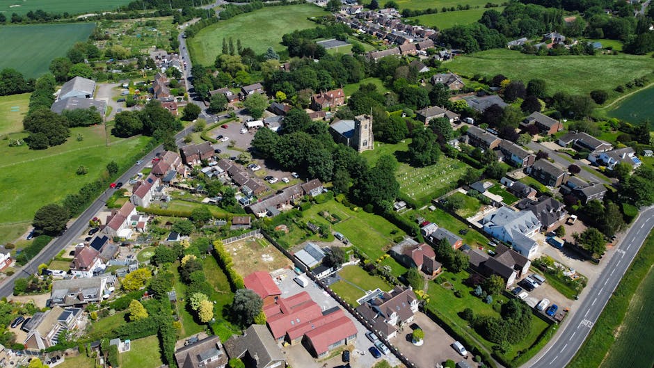 An aerial view of Pratts Bottom village showing a dense arrangement of residential houses with various roof styles, gardens, and green spaces. Several trees and patches of lawn are visible throughout the neighbourhood. A narrow road runs through the area, with parked cars along the roadside. In the foreground, there are houses with fenced gardens, some equipped with sheds or greenhouses, and others with visible patios. A church with a tall tower is situated amidst the trees towards the center of the image, indicating a local community landmark. The wider surroundings include open fields and countryside, with a mix of pathways and smaller roads connecting different parts of the village. The scene suggests a calm, well-maintained residential area suitable for home relocations, with evidence of a household moving process involving furniture, boxes, or packing materials inside the property and possibly being prepared for transportation by Man with Van Pratts Bottom as part of their removals services.
