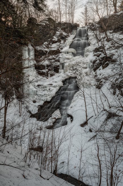 A street scene outside Pratts Bottom Village Hall during the home relocation process shows a stone-built structure with arched openings on the left side and a row of historic, beige-colored houses with pitched roofs and multiple windows on the right. In front of the houses, there is a leafless tree with sprawling branches and a well-maintained grassy area. Several vehicles, including a white van and a smaller delivery van, are parked close to the buildings, indicating the loading or unloading of furniture and packing materials. The scene is illuminated by natural daylight with a partly cloudy sky overhead, and some moving equipment such as dollies or straps may be seen in the vicinity, supporting the logistics of professional removals by Man with Van Pratts Bottom. This image captures the typical environment of a residential home relocation, where furniture transport and packing are coordinated within the local area, with the building exteriors and surrounding space clearly visible.
