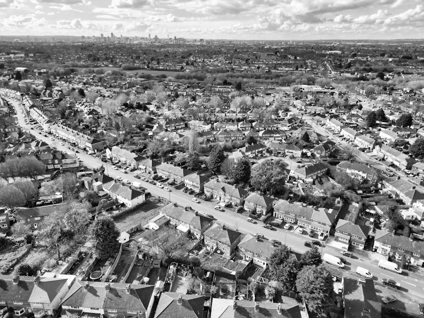 An aerial black-and-white photograph of a suburban neighbourhood showing a row of terraced houses lining a main street, with individual houses, gardens, and trees visible. The street is filled with parked cars along both sides, and some are in motion, suggesting a busy area. In the foreground, residential backyards with fences, sheds, and garden furniture can be seen, indicating the existence of a home environment. The image also captures the wider urban landscape in the distance, including larger buildings and cityscape on the horizon under a partly cloudy sky. This scenery is consistent with a home relocation or furniture transport setting, where items such as boxes, packing materials, or moving equipment might be part of the process managed by [COMPANY_NAME], facilitating quick and efficient relocations in the Pratts Bottom area, especially suitable for same-day removals requiring careful loading and transport of household furniture and belongings.