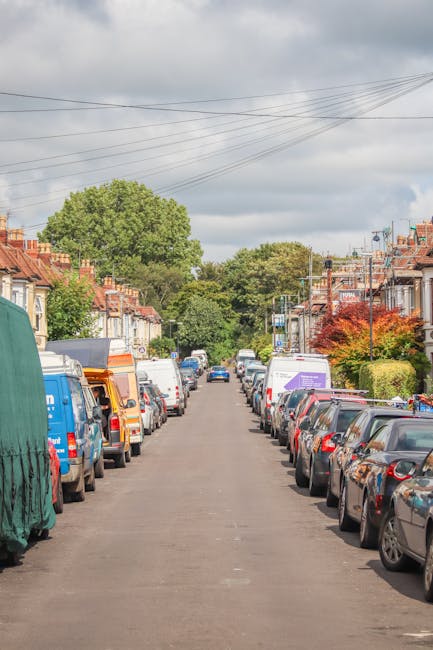 A residential street scene during daytime showing parked cars lining both sides of the road, with a row of terraced houses featuring brick facades and small front gardens. In the background, there are green trees and a partly cloudy sky. The street appears to be quiet with no visible moving vehicles or pedestrians. The image captures the typical environment for home relocation or furniture transport, relevant to house removals services such as those provided by Man with Van Pratts Bottom. The scene is well-lit with natural daylight, and the overall setting suggests a suburban neighbourhood suitable for last-minute or same-day removal services, with the focus on the residential environment and parking arrangements.