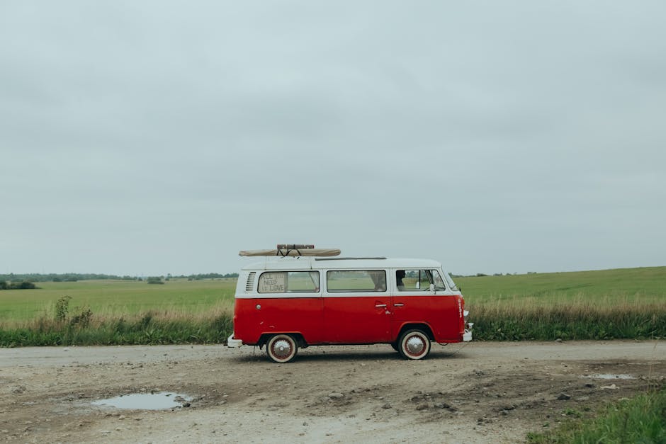 A vintage red and white Volkswagen Type 2 van parked on a rural dirt road with puddles, surrounded by green fields under a cloudy sky, with a surfboard strapped to the roof. This scene illustrates a typical setting for a home relocation or furniture transport activity, possibly associated with moving or packing services by Man with Van Pratts Bottom, as depicted on the Sevenoaks Road to Pratts Bottom page. The van's placement suggests it is either about to load or unload household goods, reflecting the logistics involved in house removals and packing during a small van-based moving process in a countryside environment.
