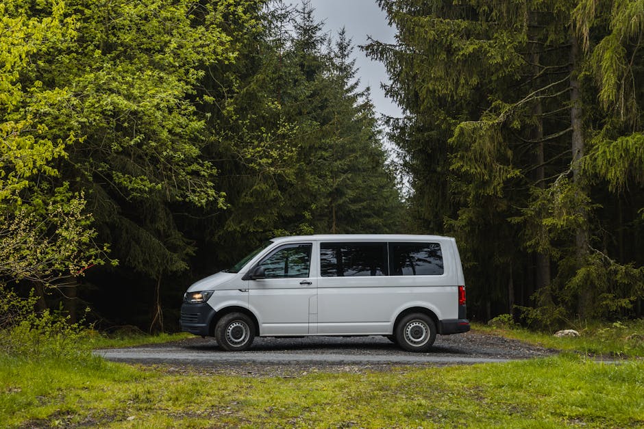 A white panel van positioned on a gravel driveway surrounded by dense green trees, with the van's side door closed and the vehicle facing left. The scene appears to be part of a home relocation or furniture transport process, as the van is parked just outside a wooded area, possibly near a house. The background features tall coniferous and broadleaf trees creating a natural, forested environment, while the foreground shows a patch of well-maintained grass. The van's windows are tinted, and the environment is lit with natural daylight, indicating an outdoor setting suitable for loading or unloading household items, boxes, or furniture as part of a professional removals service by Man with Van Pratts Bottom. The image illustrates the typical scene during the loading process of a small van used in house removals or moving logistics, emphasizing the arrangement and positioning of the vehicle for efficient home relocation.