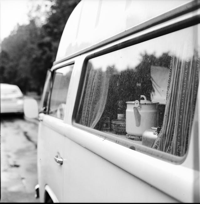 A vintage red and white Volkswagen Type 2 van parked on a rural dirt road with puddles, surrounded by green fields under a cloudy sky, with a surfboard strapped to the roof. This scene illustrates a typical setting for a home relocation or furniture transport activity, possibly associated with moving or packing services by Man with Van Pratts Bottom, as depicted on the Sevenoaks Road to Pratts Bottom page. The van's placement suggests it is either about to load or unload household goods, reflecting the logistics involved in house removals and packing during a small van-based moving process in a countryside environment.