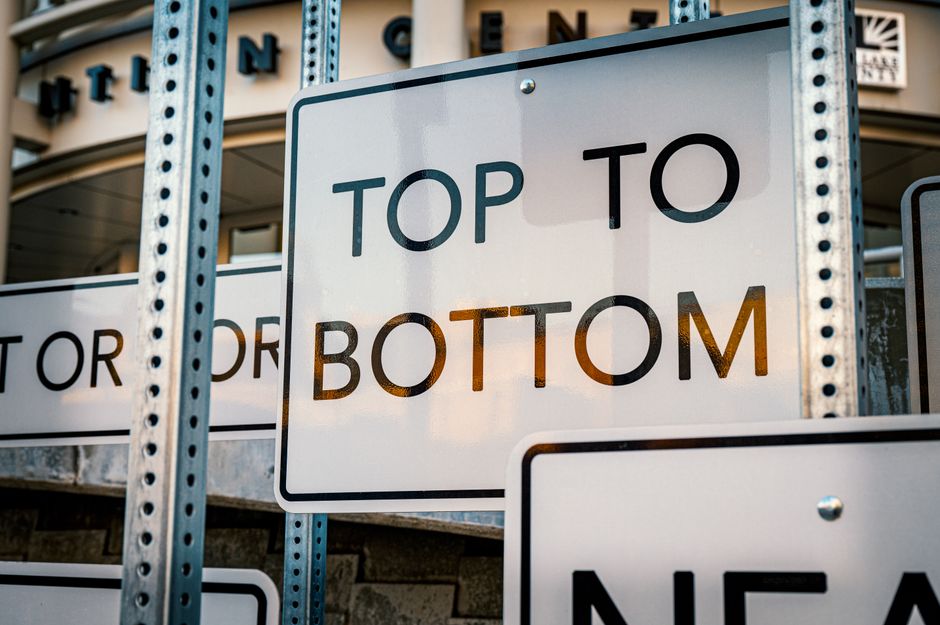 Close-up of a white, rectangular traffic sign with bold black text reading 'TOP TO BOTTOM,' mounted among a series of similar signs on a grey metal frame. The signs are situated outdoors, with the background showing a building facade with windows and part of a storefront, indicating an urban environment. The top sign is partially obscured by the foreground sign, which is sharply in focus. The lighting suggests daylight, creating shadows and reflections on the signage. This setting illustrates traffic and street management, relevant to vehicle access restrictions often encountered during house removals and home relocation processes in areas like Pratts Bottom, where adherence to street closure rules is important for smooth furniture transport and loading activities.