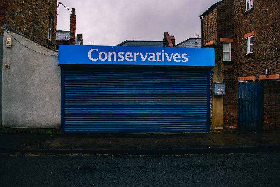 A closed blue metal roller shutter door covering a small commercial unit, with a blue sign above displaying the word 'Conservatives' in white text. The unit is situated between a white wall on the left and a brick building on the right, with a small brick extension and a blue wooden fence. A mailbox is mounted on the wall to the right of the shutter. The pavement in front of the unit is wet, suggesting recent rain, and the sky appears overcast with gray clouds. This scene likely depicts a storefront on a residential street in Pratts Bottom, appropriate for discussing house removals, furniture transport, and moving logistics as part of home relocation services offered by Man with Van Pratts Bottom.