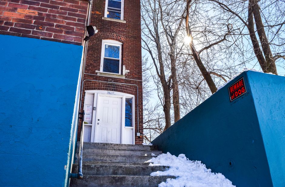 A residential brick building with a white front door accessed via a concrete staircase, flanked by bright blue painted walls on either side. The staircase has a metal handrail and some snow on the steps, indicating winter weather. Tall, leafless trees are visible in the background, with the sun shining through the branches, creating a bright glare. A small red and black warning sign reading 'BEWARE OF DOG' is affixed to the blue wall on the right side of the entrance. The scene appears to be a typical home evacuation or relocation setting, with an emphasis on the external property environment, suitable for illustrating house removals and furniture transport services provided by Man with Van Pratts Bottom as part of a house moving or packing and loading process.