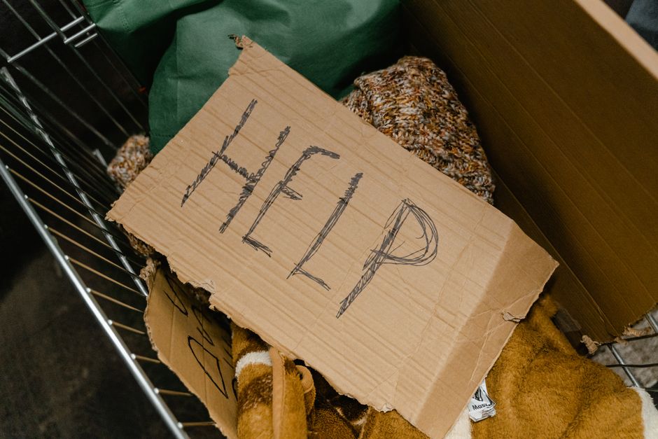 Inside a moving vehicle or storage area, a worn cardboard sign with the word 'HELP' written in black marker is placed among various packed items. Surrounding the sign are different materials used for packing, including textured fabric or blankets, multicolored knit or woven bags, and other miscellaneous packing supplies. The cardboard sign appears slightly creased and torn along the edges, indicating frequent handling or previous use. The items are situated on a surface that could be part of a moving vehicle, such as a van or truck, with metal framework visible on the left side of the image. Natural and artificial lighting illuminates the scene, highlighting the textures of the packaging materials and the handwritten message. As part of a home relocation or furniture transport process, [COMPANY_NAME] often manages such packing and loading during house removals or eviction moveouts in Pratts Bottom.