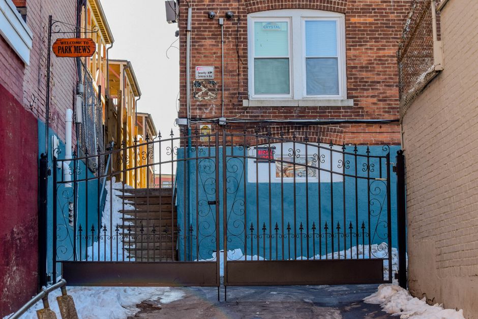 A residential brick building with a white front door accessed via a concrete staircase, flanked by bright blue painted walls on either side. The staircase has a metal handrail and some snow on the steps, indicating winter weather. Tall, leafless trees are visible in the background, with the sun shining through the branches, creating a bright glare. A small red and black warning sign reading 'BEWARE OF DOG' is affixed to the blue wall on the right side of the entrance. The scene appears to be a typical home evacuation or relocation setting, with an emphasis on the external property environment, suitable for illustrating house removals and furniture transport services provided by Man with Van Pratts Bottom as part of a house moving or packing and loading process.